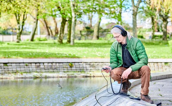 Person in a green jacket and headphones crouching by a park pond, using electronic equipment with cables extending into the water. Trees, grass, and a stone wall line the background.